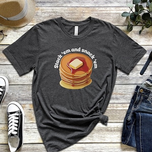 Puede incluir: Una camiseta gris oscuro jaspeada con un gráfico de una pila de panqueques con mantequilla y jarabe. El texto en la camiseta dice "Stack 'em and snack 'em".