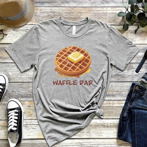 K&ouml;nnte beinhalten: Ein graues T-Shirt mit einem Cartoon-Waffel mit Butter und Sirup. Der Text "Waffle Dad" ist unter der Waffel gedruckt.