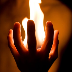 Op de afbeelding: Een hand met gespreide vingers, die een vlam in de palm houdt. De vlam is helder oranje en geel, en de hand is gesilhouetteerd tegen het licht.