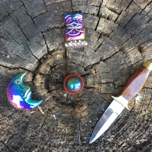 Puede incluir: Un trío de objetos iridiscentes, de colores arcoíris, sobre una superficie de madera. Una luna creciente, una pequeña estatua y un pequeño cuchillo con mango de madera.