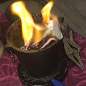 Puede incluir: Un crisol de metal negro con una base de madera se encuentra sobre una superficie morada. El crisol está lleno de papel en llamas y se ven llamas.