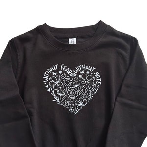 May include: Black sweatshirt with a white heart design that says "Without Fear Without Hate" with floral accents.