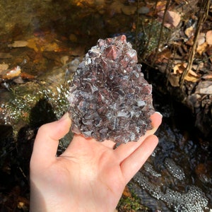 May include: A hand holds a cluster of dark red-brown crystals, showcasing a layered, sparkling texture. The crystals are set against a backdrop of a flowing stream and natural foliage, creating a contrast of textures and colours.