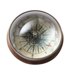 May include: A vintage compass with a clear, domed glass cover. The compass face is beige with black directional markings and numbers. The outer casing is bronze-coloured. The compass needle points to the southeast.
