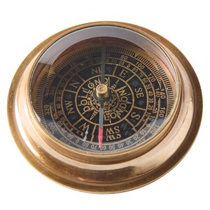 May include: A vintage brass compass with a clear glass top. The compass face is black with gold lettering indicating directions and degrees. The needle points south. The compass is a navigational tool.
