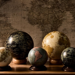 May include: Several antique globes in different sizes and colours, including black, cream, and blue, each on a wooden base. They are arranged on a wooden surface against a backdrop of a vintage world map.
