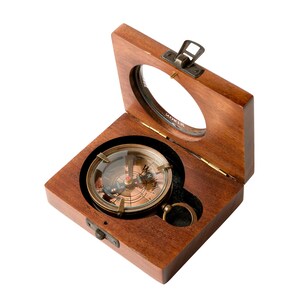 May include: A round brass compass with a glass cover and a metal ring pull, set inside a square wooden box. The box features a hinged lid with a circular glass window, a metal latch closure, and a dark brown finish with visible wood grain.