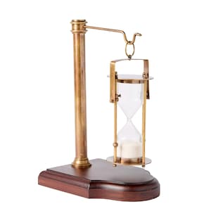 May include: A brass hourglass suspended from a horizontal arm attached to a vertical brass post, all mounted on a dark brown wooden base. The hourglass features clear glass bulbs containing white sand, held within a brass frame with circular ends.