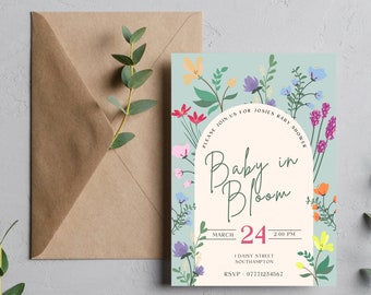 May include: A baby shower invitation with a floral design. The invitation features the text "Baby in Bloom" in a script font, surrounded by colorful flowers. A brown envelope and a sprig of greenery are also included.