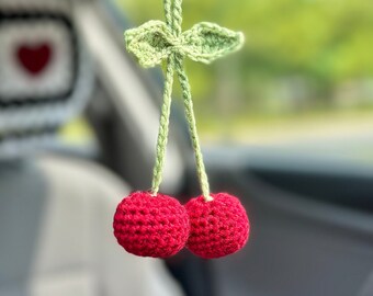 May include: A pair of red crocheted cherries with green crocheted leaves hanging from a rearview mirror in a car.