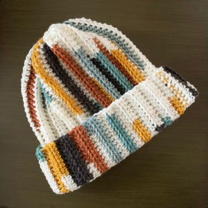 May include: A handmade crochet beanie in a ribbed pattern. The hat features vertical stripes in cream, mustard yellow, brown, teal, and black. The brim is folded up, showcasing the striped design. The beanie is designed for warmth and style.