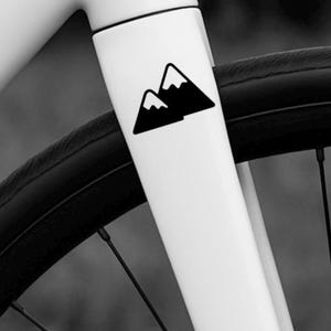 May include: Close-up of a white bicycle frame and front fork with a black mountain logo. The black tire and spokes are visible, along with the disc brake. The bike is designed for road cycling.