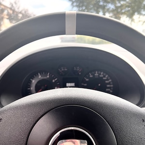 May include: A close-up view of a black steering wheel with a gray stripe at the top. The dashboard is visible in the background, with gauges and the word "DRIVE" displayed. The steering wheel has a textured grip.