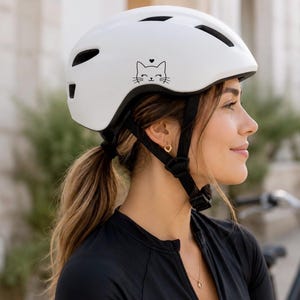 Pode incluir: Capacete de bicicleta branco com um desenho de rosto de gato preto e um coração. O capacete tem correias pretas ajustáveis. Design moderno e seguro para ciclismo.