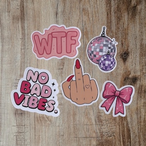 Op de afbeelding: Een set van vijf stickers met een roze sticker met de tekst WTF in bubbelletters, een roze en paarse discobal, een hand die een middelvinger opsteekt met rode nagels en een hart-tattoo, een roze strik en een sticker met de tekst NO BAD VIBES.