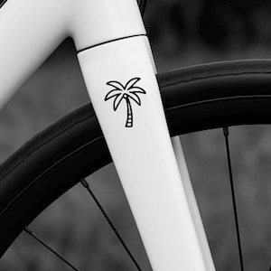 Pode incluir: Imagem a preto e branco de uma estrutura de bicicleta branca com um desenho de palmeira preta. A palmeira é um desenho de linha simples. A roda da bicicleta é visível no fundo.