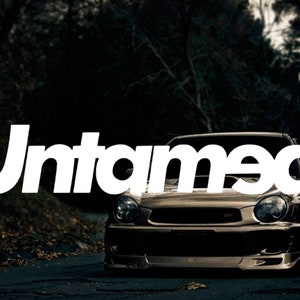 Könnte beinhalten: Ein dunkelgraues Auto mit dem Wort "Untamed" in weißen Buchstaben auf der Front. Das Auto steht auf einer asphaltierten Straße vor einem dunklen Wald.