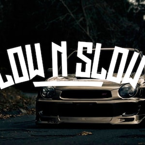 Puede incluir: Un coche negro con las palabras "LOW N SLOW" en letras blancas en el parabrisas delantero. El coche está aparcado en una carretera de asfalto oscura.