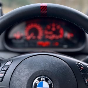 Könnte beinhalten: Nahaufnahme eines schwarzen Lenkrads mit einem rot-schwarzen Streifen oben. Das Armaturenbrett hinter dem Lenkrad zeigt rot beleuchtete Anzeigen. Das BMW-Logo ist in der Mitte des Lenkrads zu sehen.