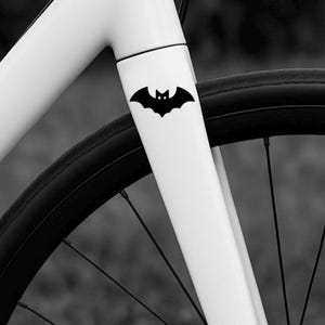 May include: A black bat decal is affixed to the white frame of a bicycle. The bat has a stylized design with pointed ears and a small, centered face. The bicycle wheel and spokes are visible in the background.