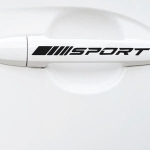 May include: A white car door handle with a black "SPORT" decal. The decal includes the word "SPORT" in a bold, sans-serif font, with a stylized graphic above the word.