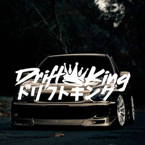 Puede incluir: Un coche negro con una pegatina blanca en el capó que dice "Drift King" con una corona sobre la palabra "Drift". La pegatina también tiene texto japonés que dice "ドリフトキング".