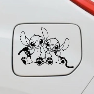Op de afbeelding: Zwart-witte sticker met twee stripfiguren, Stitch, uit de animatiefilm Lilo & Stitch. De figuren zitten samen, een houdt een gereedschap vast. De sticker is aangebracht op een wit oppervlak.
