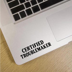 Puede incluir: Un ordenador portátil blanco con un teclado negro y una pegatina que dice "CERTIFIED TROUBLEMAKER" en letras negras en negrita. El portátil está sobre una superficie de madera de color claro.