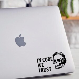 Op de afbeelding: Een witte laptop met een zwarte sticker met de tekst "IN CODE WE TRUST" naast een schedel met een koptelefoon. De laptop heeft een zilveren Apple-logo.