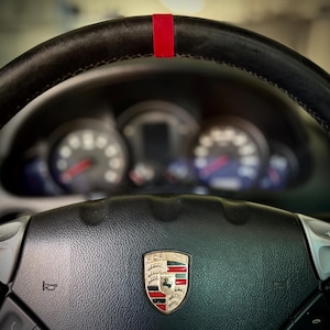 May include: Close-up of a black leather steering wheel with a red stripe at the top. The Porsche emblem is centered on the wheel. Gauges are visible in the background.
