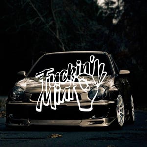 Può includere: Un'auto nera con un adesivo bianco sul parabrezza che dice "Fuckin' Minty".