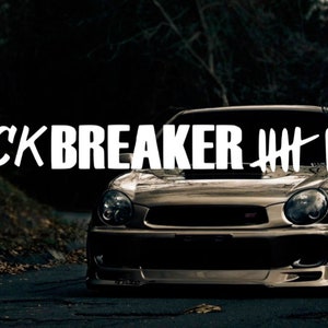 Op de afbeelding: Een zilveren Subaru Impreza met een witte "Neckbreaker" sticker op de voorruit. De auto staat geparkeerd op een geasfalteerde weg met bomen op de achtergrond.