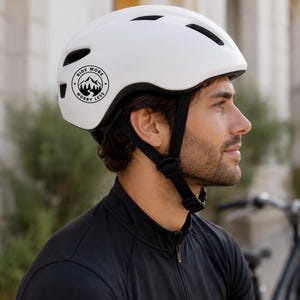 Op de afbeelding: Witte fietshelm met zwarte bandjes en ventilatieopeningen. Een zwart-witte cirkelvormige afbeelding aan de zijkant leest "Ride More Worry Less" met een berg- en boomontwerp. De helm is ontworpen voor veiligheid en buitenactiviteiten.
