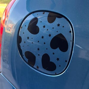 Puede incluir: Una tapa de gasolina de coche azul con una pegatina de vinilo negro con un patrón repetido de corazones y estrellas negras.