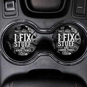 Op de afbeelding: Twee zwarte auto-onderzetters met een wit ontwerp met de tekst "That's what I do I fix stuff and I know things".