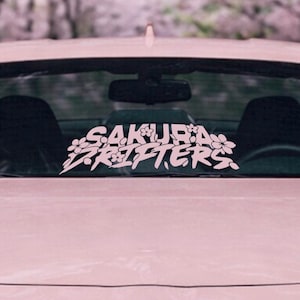 Puede incluir: Calcomanía de vinilo blanca para coche con el texto "SAKURA DRIFTERS" en una fuente estilizada, adornada con ilustraciones de flores de cerezo. La calcomanía se muestra en la ventana trasera de un coche rosa, con un fondo borroso de árboles.