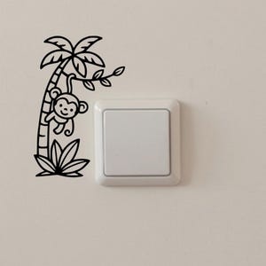 Puede incluir: Adhesivo de pared en blanco y negro con un mono de dibujos animados colgado de una palmera. El diseño incluye un contorno detallado del árbol, las hojas y un mono sonriente. La calcomanía se coloca cerca de un interruptor de luz blanco.
