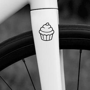 Peut inclure: Image en noir et blanc d'un vélo. Un autocollant de cupcake noir est sur le cadre blanc du vélo. Le pneu du vélo est noir. L'autocollant de cupcake est un simple dessin au trait.