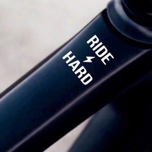 Peut inclure: Gros plan sur un cadre de vélo bleu foncé avec les mots "RIDE HARD" en blanc et un symbole d'éclair. Le cadre a une finition lisse et mate.