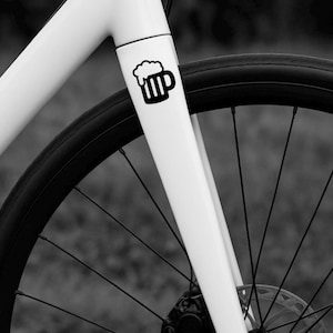 Op de afbeelding: Close-up van een wit fietsframe met een zwarte bierpulafbeelding aan de voorkant. De zwarte band en spaken van het voorwiel zijn zichtbaar, tegen een vage achtergrond.