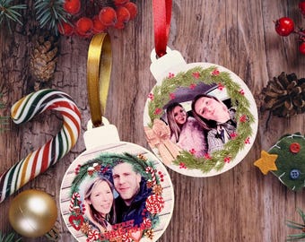 May include: Two wooden Christmas ornaments with photos inside, surrounded by a wreath design. One has a gold ribbon, the other a red ribbon. Festive holiday decor includes candy canes, pine needles, and berries.