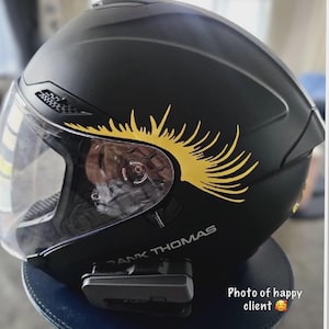 Op de afbeelding: Een matzwarte motorhelm met een helder vizier en een gele afbeelding van lange wimpers boven het vizier. De helm is voorzien van de tekst FRANK THOMAS in witte letters en heeft een zwart communicatieapparaat aan de zijkant bevestigd.