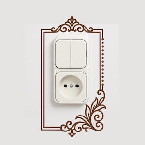 Op de afbeelding: Een witte stopcontact- en lichtschakelaarcombinatie met een decoratief bruin frame. Het frame heeft sierlijke bloemen- en krulontwerpen, die een vleugje elegantie aan de muur toevoegen.