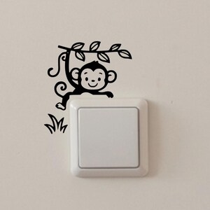 Może przedstawiać: Czarna naklejka z uśmiechniętą małpką zwisającą z gałęzi z liśćmi. Małpka jest umieszczona nad białym włącznikiem światła, z detalami przypominającymi trawę po lewej stronie. Ta naklejka jest odpowiednia do pokoju dziecięcego.