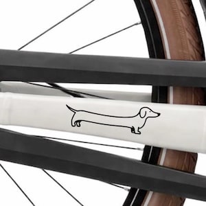 Op de afbeelding: Zwart-wit illustratie van een teckel op een witte fietsframebeschermer. De hond is afgebeeld in een eenvoudige, minimalistische stijl. Het fietswiel en frame zijn zichtbaar op de achtergrond.