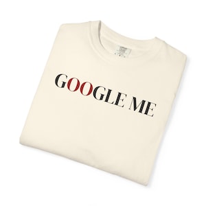 May include: Cream-colored t-shirt with the phrase "GOOGLE ME" printed in black and red letters. The shirt is folded neatly, showcasing the design prominently. The tag is visible at the top.