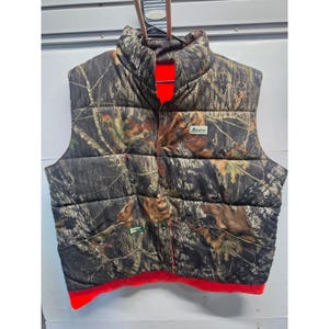 Puede incluir: Un chaleco acolchado sin mangas con un patrón de camuflaje de corteza de árbol y hojas en tonos marrón, negro y beige. El chaleco tiene un cuello alto, cierre frontal con botones y un forro interior de color naranja brillante visible.