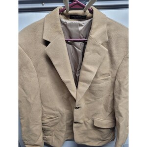 May include: A tan blazer with a notched lapel, two front pockets, and a single-button closure. The interior lining is a lighter tan color. The blazer is hanging on a pink hanger.