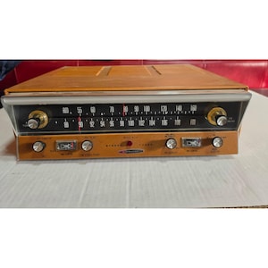 May include: A Heathkit model AJ-41 stereo tuner with a tan casing and a black front panel. The device features a horizontal radio frequency display for AM and FM bands, multiple silver control knobs, and two small rectangular signal meters.