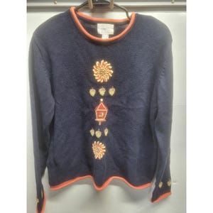 May include: A navy blue long-sleeve sweater with an orange trim at the crew neckline, cuffs, and hem. The front features a vertical embroidered design of two sunflowers, several small acorns, and a central red birdhouse with a yellow accent.